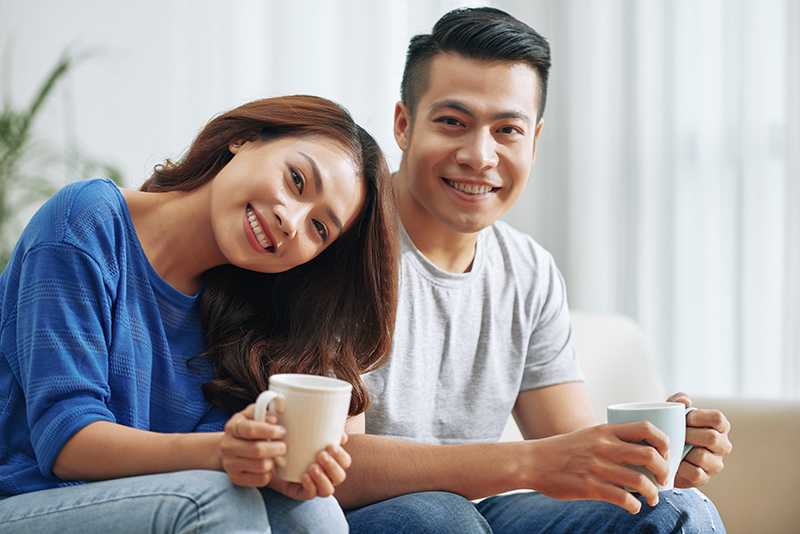 happy-asian-couple-sitting-couch-home-with-tea-mugs-smiling happy-asian-couple-sitting-couch-home-with-tea-mugs-smiling